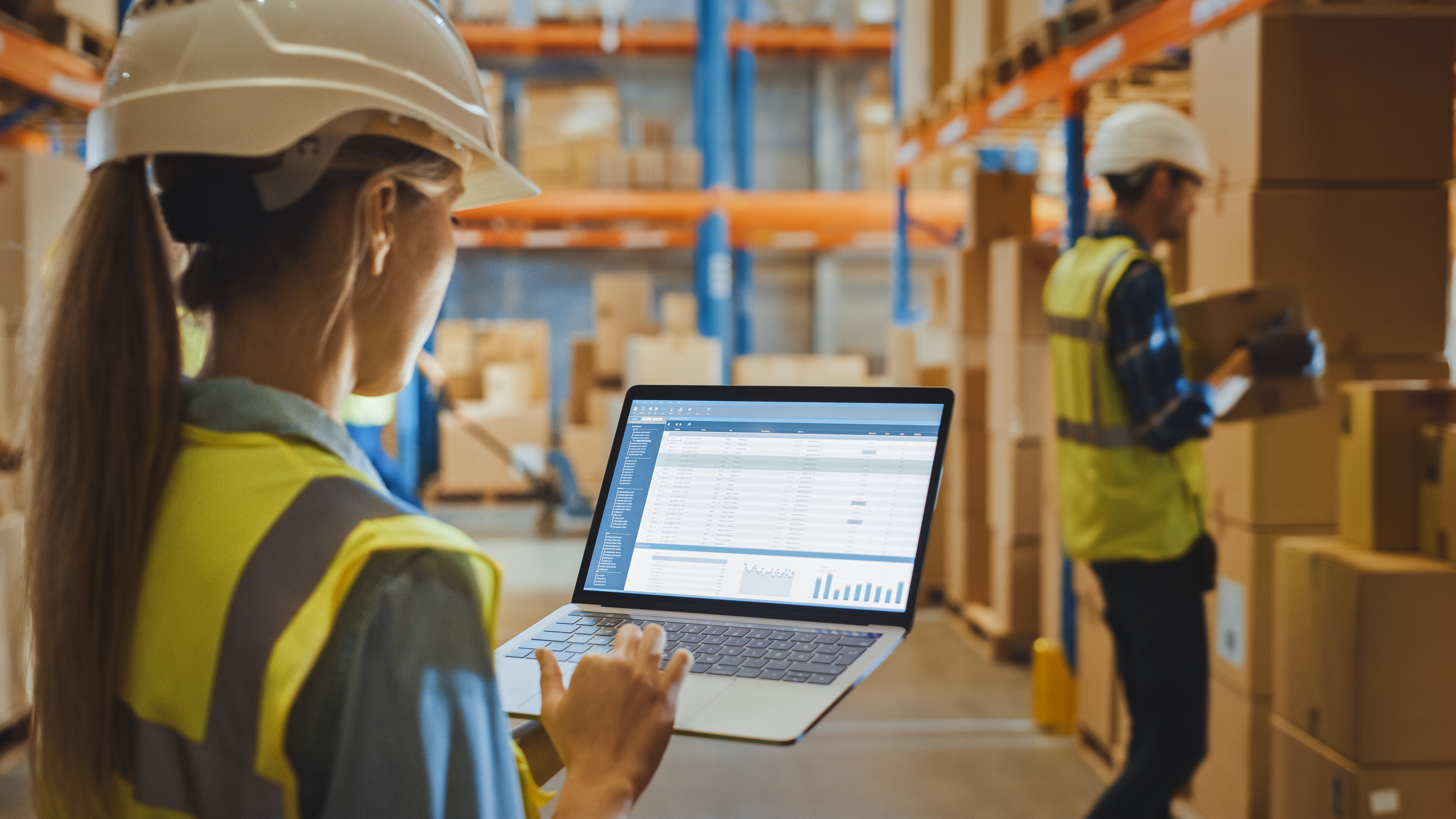 Kvindelig lagerarbejder med sikkerhedshjelm holder en bærbar og gennemgår en rapport om Warehouse Profitability.
